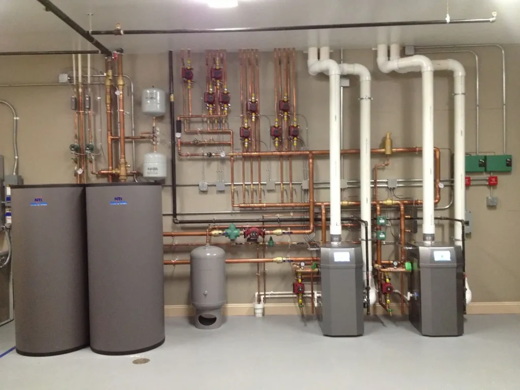 Large mechanical room with two boilers, storage tanks, circulation pumps, copper piping, and white vent pipes.