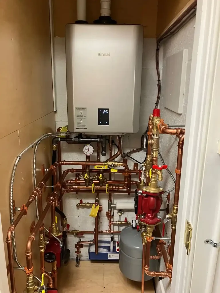 Wall-mounted Rinnai unit connected to copper piping, circulation pumps, valves, gauges, and an expansion tank.