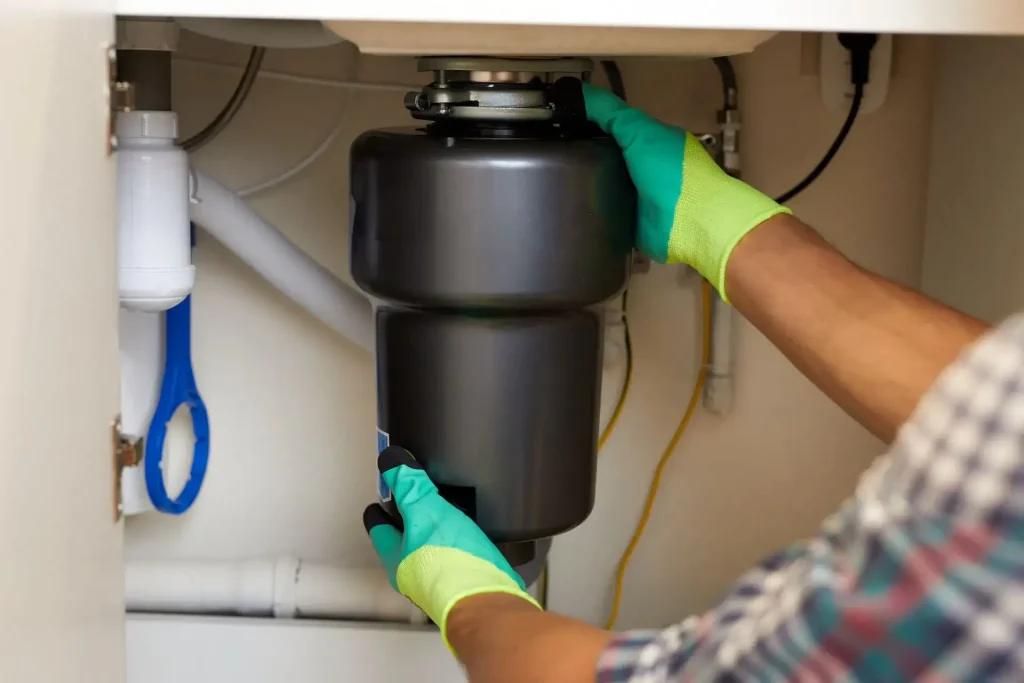 Gloved technician installing a black garbage disposal beneath a kitchen sink.