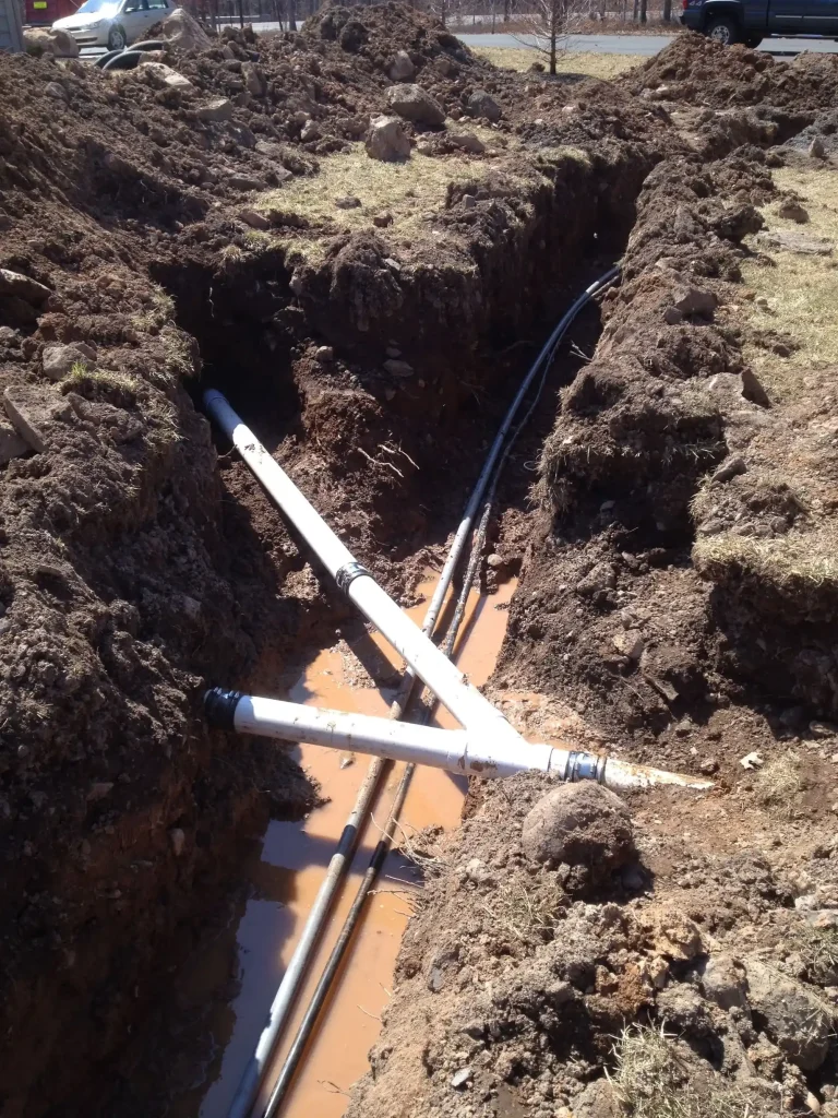 White sewer pipe and utility lines exposed in a deep, muddy trench surrounded by excavated soil.