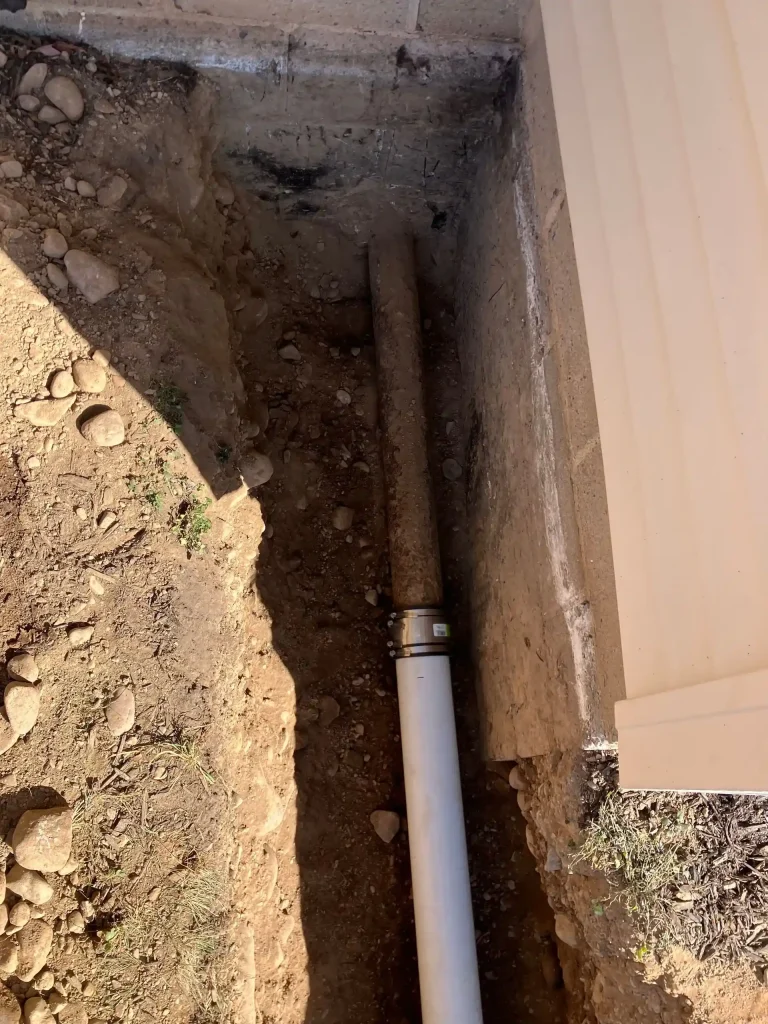 Excavated trench beside a building exposing an old underground pipe connected to a new white PVC sewer line.