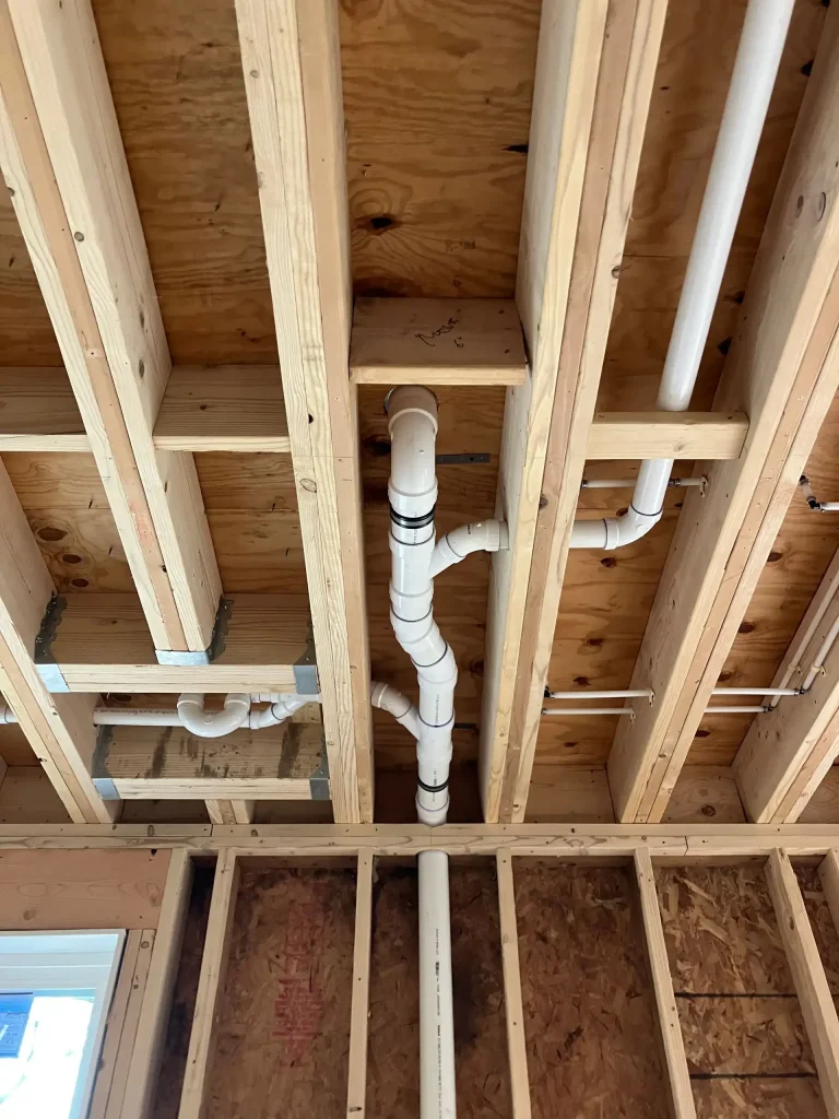 White PVC drain and vent pipes installed between exposed wooden ceiling and wall framing.