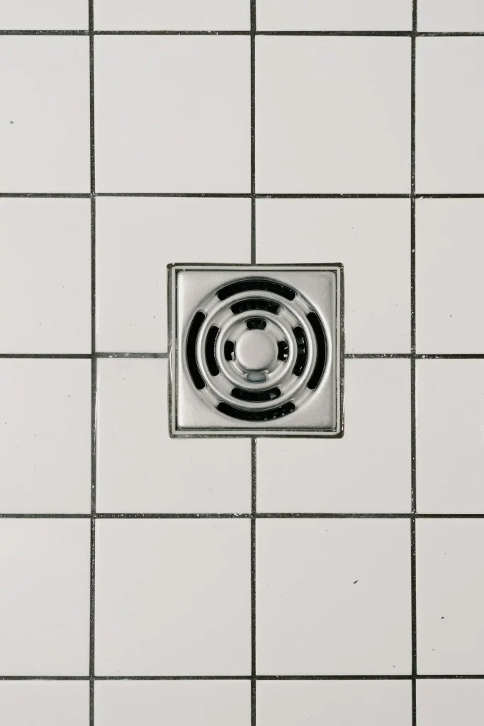 Square stainless-steel floor drain installed among white tiles with dark grout.