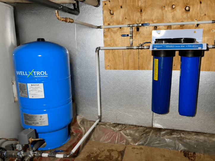 Blue well pressure tank connected to a two-stage whole-house water filtration system and supply piping.